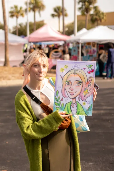 A blonde person wearing elf ears holding up a stylized self-portrait of themselves.